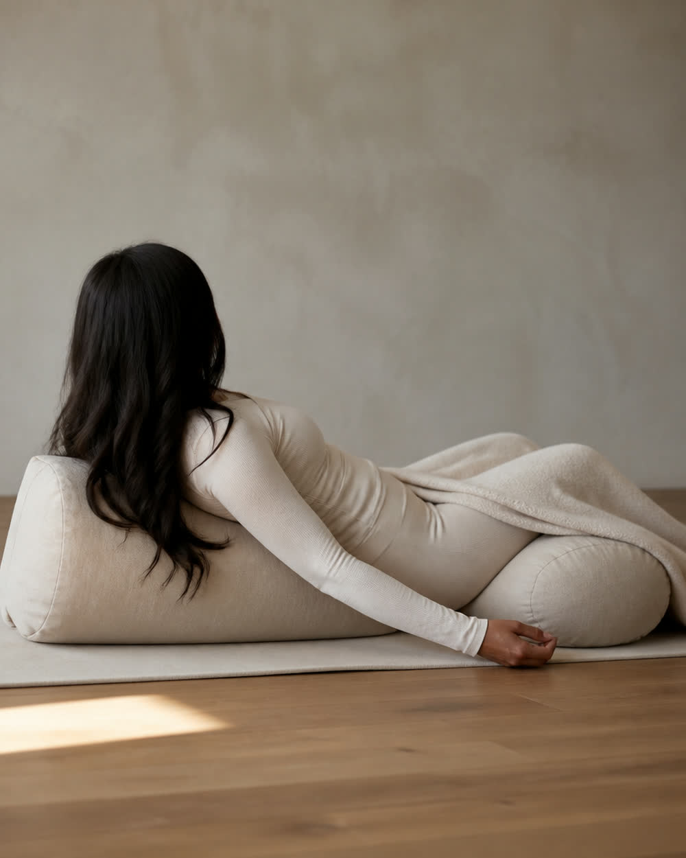 A reclined shape fully supported over a bolster, a folded blanket across the legs, on a quiet wooden floor.
